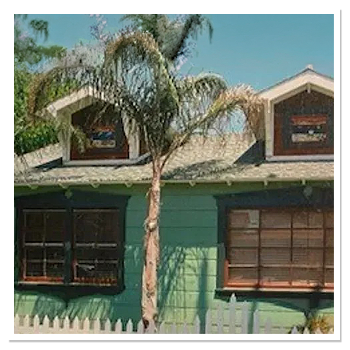 Green Craftsman house with two front dormers and dark window trim behind a palm tree and picket fence