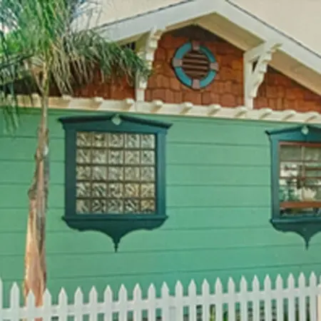 Green house with a glass-block window, porthole vent and cedar-shingle gable behind a picket fence